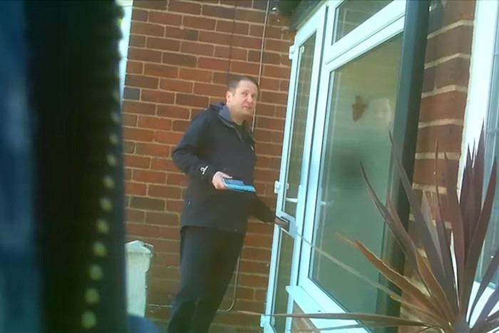 Alan Feeney stands outside a brick house holding campaign leaflets, reaching for the door handle.