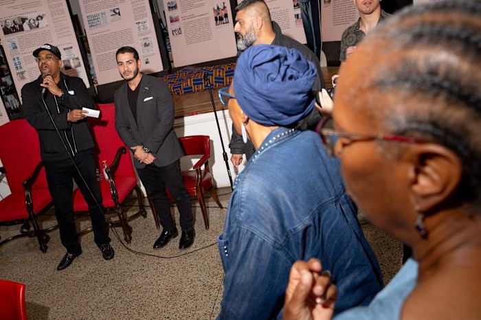 A group of people stands and listens as a man with a microphone speaks at an indoor gathering, with informational posters displayed behind them.