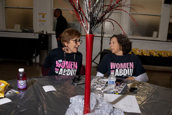 Two women wearing “Women for Abdul” shirts sit at a decorated table, conversing at an indoor event.