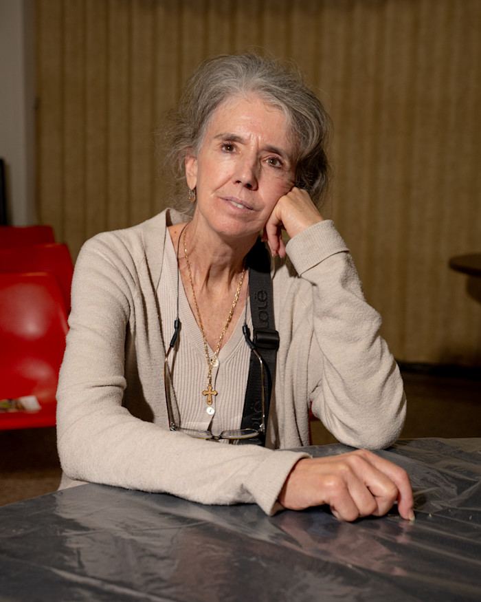 Marianne Livezey seated at a table, resting her head on her hand, with red chairs and a curtain in the background.