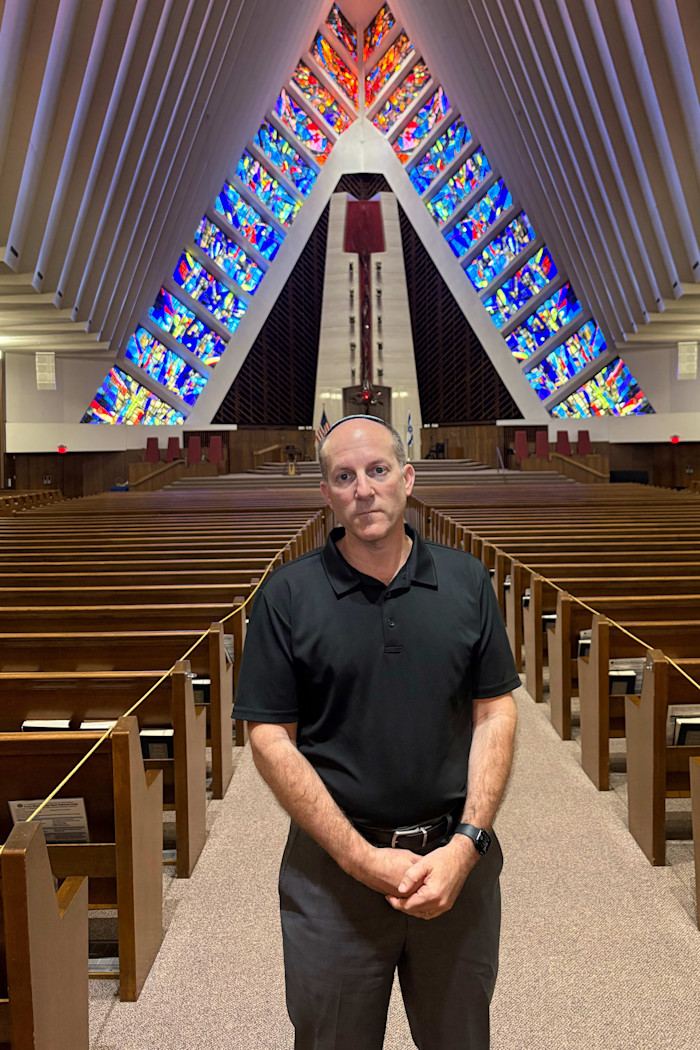 Rabbi Aaron Starr stands in the sanctuary of Community Shaarey Zedek, with rows of pews and colorful stained glass windows behind him.
