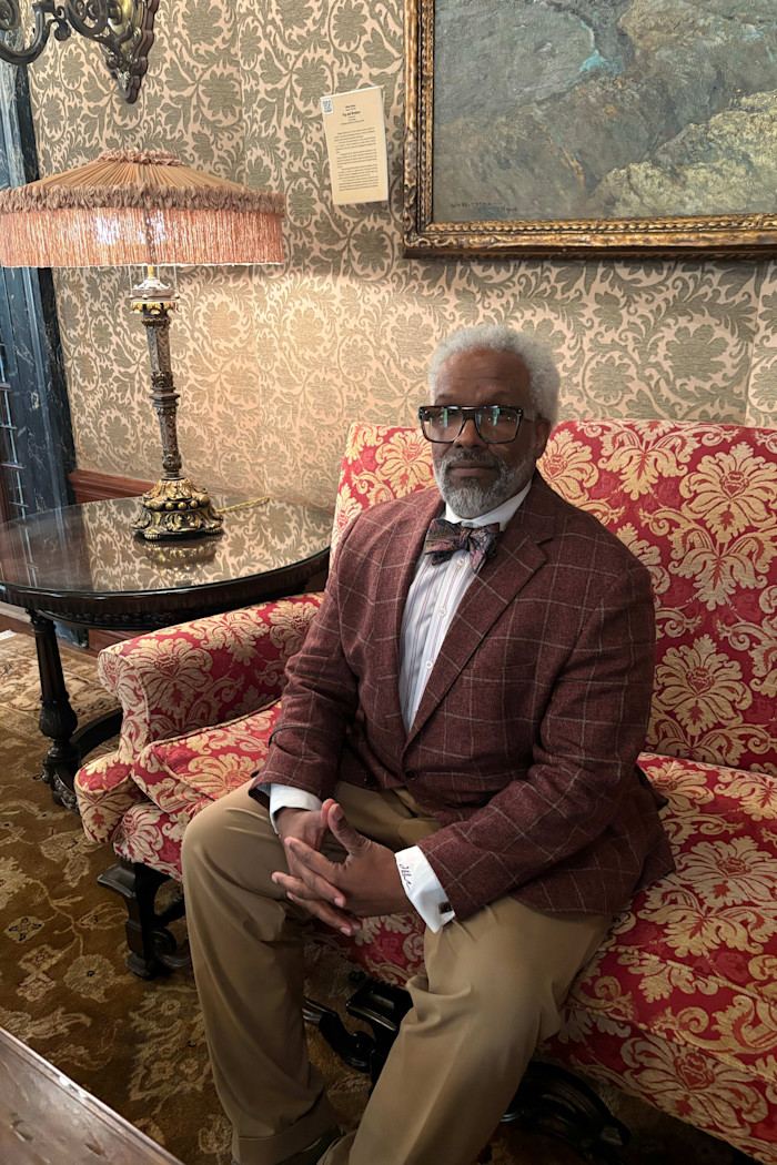 Todd Perkins sits on a patterned red and gold sofa, wearing a plaid blazer, striped shirt, and bow tie.