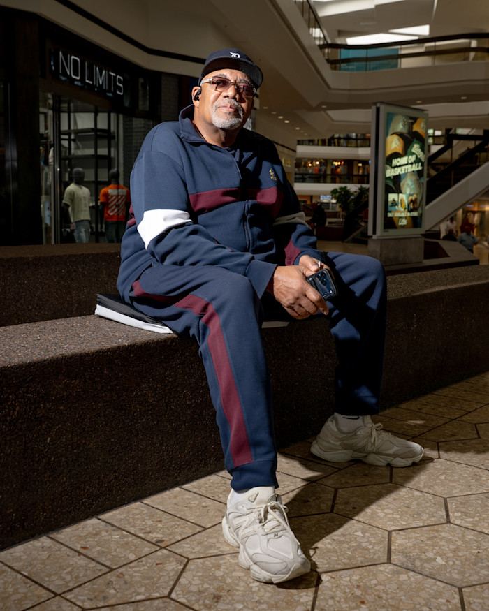Stu Jackson sits on a bench at Fairlane Mall, wearing a tracksuit, cap, and sunglasses, holding a phone.