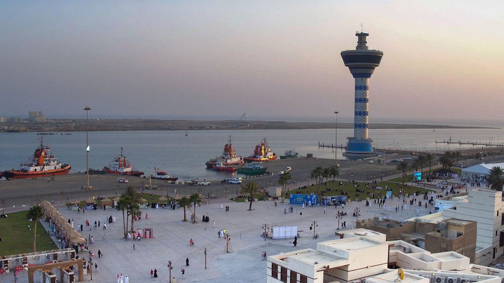 A waterfront scene at Yanbu port shows several docked boats, a tall blue and white control tower, and a plaza with crowds of people.