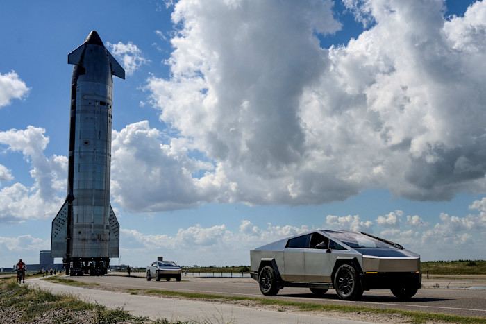 Tesla Cybertrucks drive ahead of the SpaceX Starship spacecraft as it is transported towards a launchpad under a partly cloudy sky.