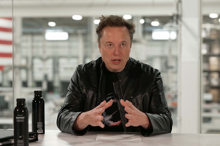 Elon Musk sits at a table speaking, gesturing with his hands during the SpaceX results announcement.