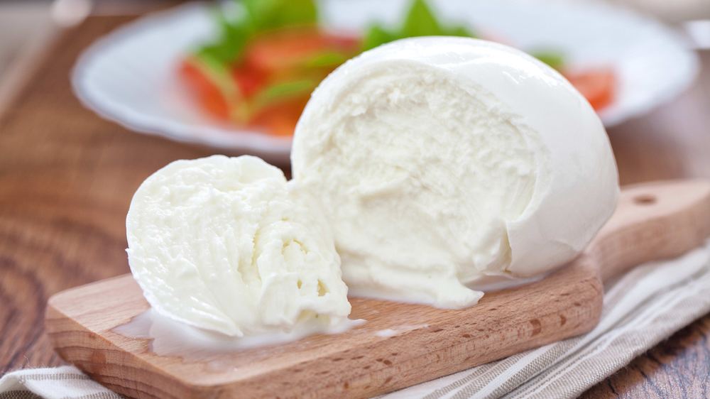 A ball of buffalo mozzarella cheese, partially sliced, sits on a wooden cutting board with a plate of tomatoes and greens in the background.