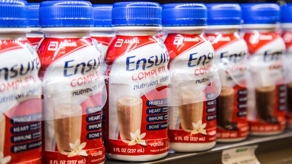 Bottles of Ensure Complete nutrition shakes with blue caps arranged in a row on a grocery store shelf.